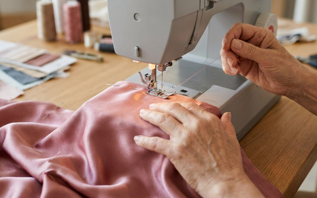 Mains d'une personne âgée cousant un tissu en soie rose avec une machine à coudre sur une table en bois.